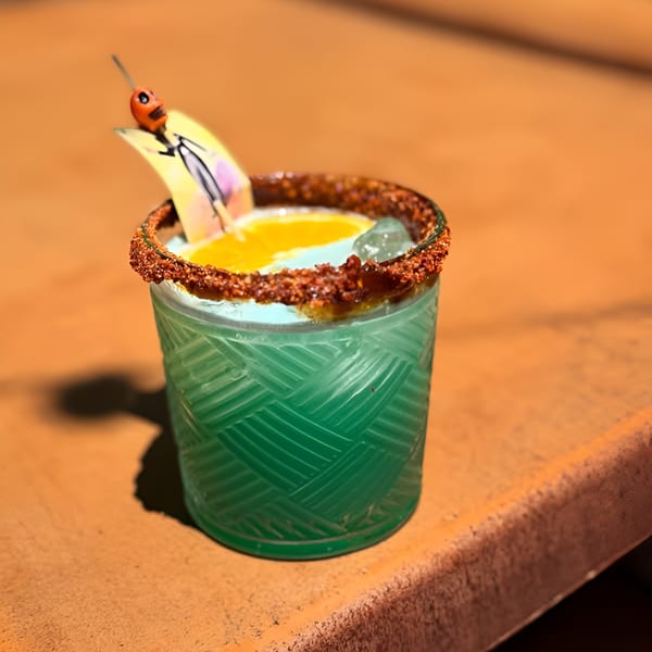 A vibrant green cocktail with a chili-lime rim, orange slice, and a decorative pick on a rustic surface.